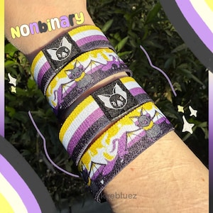 Puede incluir: Brazaletes no binarios con un diseño de murciélago. Las pulseras presentan los colores de la bandera del orgullo no binario: amarillo, blanco, morado y negro. El texto "Nonbinary" es visible en la pulsera superior.