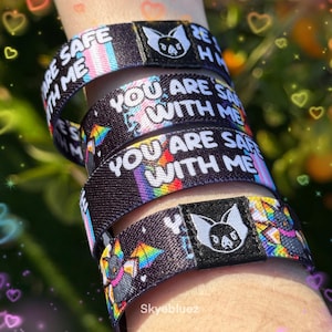 May include: A stack of black and rainbow wristbands with the text "YOU ARE SAFE WITH ME" and a bat logo. One wristband has the text "ARE SAFE WITH ME". The wristbands are on a wrist.