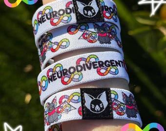 May include: White wristbands with the word "NEURODIVERGENT" and a colorful infinity symbol design. Each band features a black square with a bat logo. The wristbands are stacked on top of each other.