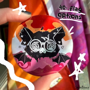 May include: A round button with a bat design wearing glasses, in black and white, set against a background of orange, red, and pink. The text "40+ flag options" is written in white. The button is held by a person.