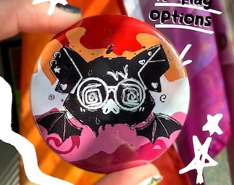 May include: A round button with a bat design wearing glasses, in black and white, set against a background of orange, red, and pink. The text "40+ flag options" is written in white. The button is held by a person.