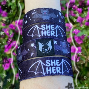May include: A set of three black elastic wristbands featuring a repeating pattern of small grey bats and white stars. Two bands display the text SHE HER in white letters framed by bat wings. One band features a central black square patch.