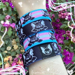 May include: A set of three elastic wristbands featuring a repeating bat graphic. Two bands have a light blue and pink striped pattern, while the third is solid black. A small black fabric tag with a white bat icon is attached to the black band.