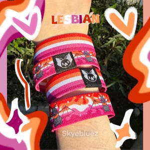 May include: A stack of elastic wristbands with the lesbian pride flag colors: pink, orange, and white. Each band features a bat graphic on a black label. The word "LESBIAN" is visible in the image.