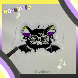 May include: A die-cut sticker featuring a black bat illustration with spiral-patterned glasses. The bat's ears display the yellow, white, purple, and black nonbinary pride flag colors. The word "nonbinary" is printed in yellow and purple above the bat.