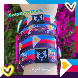 May include: A stack of colorful wristbands with a bat logo and the word "Polyamorous". The bands feature a design with red, white, blue, and yellow colors. The text "Skyebluez" is also visible.
