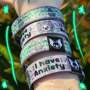 May include: Gray wristbands with a bat and wing design, and the text "I have Anxiety". The wristbands have a green and black color scheme. The wristbands are stacked on top of each other.