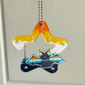 May include: A star-shaped acrylic charm hanging from a silver ball chain. The charm features a gradient of orange, yellow, and white at the top, with a dark blue base. A cartoon bat with wings colored in yellow, white, and blue is centered.