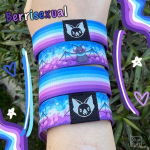May include: Wristbands in blue, purple, and white, with a bat illustration and the text "Berrisexual". The bands are stacked on top of each other. The design is a vibrant mix of colours.