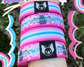 May include: Pink and blue striped wristbands with bat and bat-wing designs. The wristbands have a black label with a bat logo. The word "Femboy" is written in pink and blue on the top left.