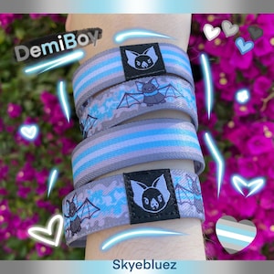 Puede incluir: Pulseras a rayas grises y azules con gráficos de murciélagos. Cada pulsera presenta un parche cuadrado negro con un diseño de murciélago. Los textos "DemiBoy" y "Skyebluez" son visibles. Las pulseras están apiladas.