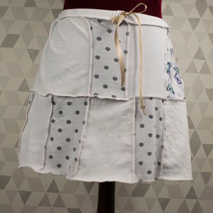 May include: A white skirt made from different pieces of fabric with a floral pattern and a small butterfly pattern. The skirt has a gold ribbon tied at the waist.