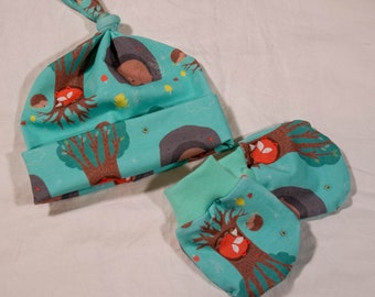 May include: A baby hat and matching mittens set. The set has a teal background with a repeating pattern of trees, foxes, hedgehogs, and mushrooms. The hat is knotted at the top, and the mittens have a teal cuff.