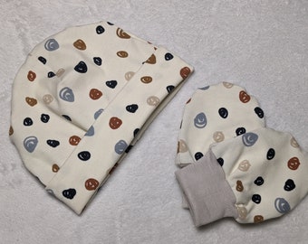 May include: A set of baby accessories in a cream colour with a brown, black and blue abstract pattern. The set includes a beanie and a pair of mittens.