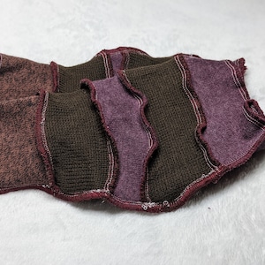 May include: A multi-coloured knitted fabric with brown, purple and maroon stripes. The fabric has a frayed edge and is folded in half.