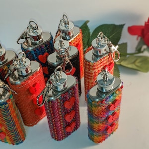 May include: A group of ten small, silver hip flasks with red heart-shaped buttons. The flasks are covered in colourful patterned fabric.