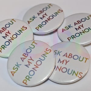 May include: Five white round buttons with the text "Ask About My Pronouns" in rainbow colours. The buttons are arranged in a circle.