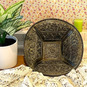 May include: A round, dark-toned metal bowl featuring a square center with a sunburst pattern and four surrounding sections decorated with embossed floral and leaf motifs. The bowl has a raised, textured rim and a deep, sloped interior design.