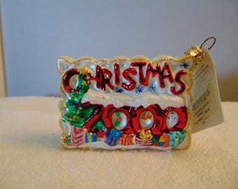 May include: A rectangular glass Christmas ornament featuring the red text CHRISTMAS above the year 2000 in large red numerals. A green Christmas tree and colorful gift boxes are at the base, with white glittery snow accents and a gold hanging loop.