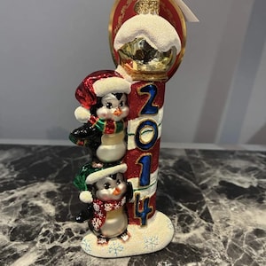 May include: A glass Christmas ornament featuring two penguins stacked vertically next to a red and white striped pole with the blue text 2014. The penguins wear festive hats and scarves, and the base and top are covered in white glittery snow.