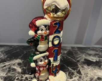 May include: A glass Christmas ornament featuring two penguins stacked vertically next to a red and white striped pole with the blue text 2014. The penguins wear festive hats and scarves, and the base and top are covered in white glittery snow.