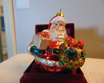 May include: A glass Santa Claus ornament featuring a red suit with white trim, a black belt, and a gold buckle. Santa holds a striped gift box in one hand and a green wreath with red berries and the gold text 2009 in the other hand.