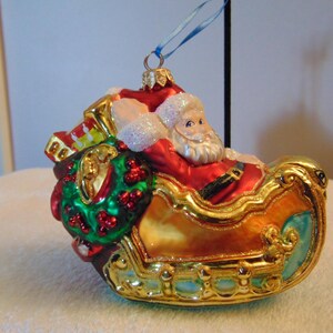 Könnte beinhalten: Ein Weihnachtsschmuck aus Glas in Form eines Schlittens mit dem Weihnachtsmann. Der Schlitten ist goldfarben mit blauen Akzenten und der Weihnachtsmann trägt einen roten Anzug. Ein grüner Kranz mit roten Beeren ist hinten befestigt.