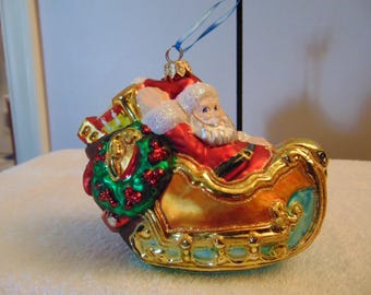 May include: A glass Christmas ornament shaped like a sleigh carrying Santa Claus. The sleigh is gold with blue accents, and Santa wears a red suit. A green wreath with red berries is attached to the back, and a blue ribbon is tied to the top.