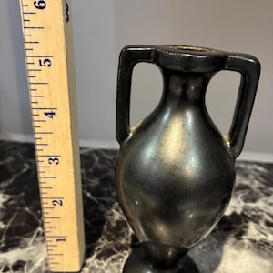 Pu&ograve; includere: Un piccolo vaso nero in stile anfora con due manici laterali, posizionato accanto a un righello di legno che mostra che l'oggetto &egrave; alto circa 14 cm. Il vaso ha una finitura scura e liscia e una base circolare.