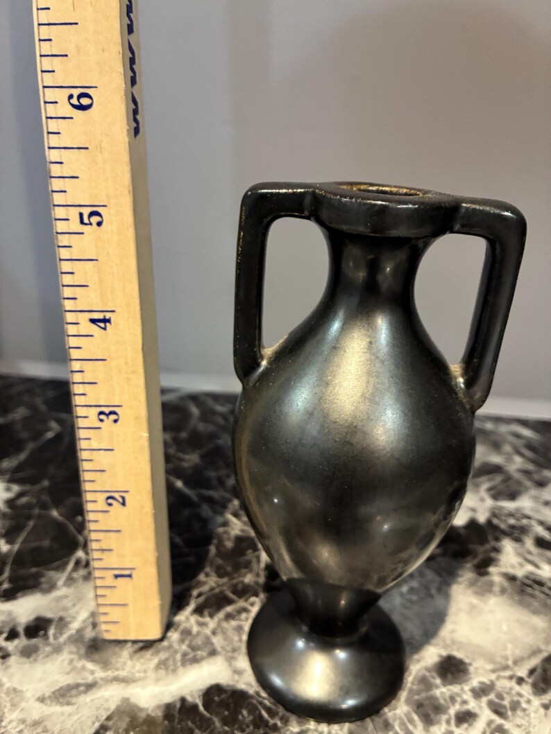 Pu&ograve; includere: Un piccolo vaso nero in stile anfora con due manici laterali, posizionato accanto a un righello di legno che mostra che l'oggetto &egrave; alto circa 14 cm. Il vaso ha una finitura scura e liscia e una base circolare.