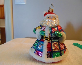 May include: A glass Santa Claus ornament featuring a patchwork coat with various colorful patterns, including red awareness ribbons, green polka dots, and candy cane stripes. The figure has a white beard, a red hat, and white trim.