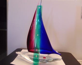 May include: A glass sailboat sculpture featuring a deep red sail and a vibrant blue sail, set upon a clear glass base. A green plastic ruler is positioned vertically against the sails, showing a height measurement of approximately 9 inches.