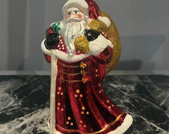 May include: A glass Santa Claus ornament with a shiny red coat, white trim, and a gold glittery sack. A wooden ruler is positioned behind the ornament, showing a height of approximately 6 inches. The figure holds a staff and wears black gloves.