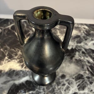 Pu&ograve; includere: Un piccolo vaso in ceramica nera con corpo arrotondato, collo stretto e due manici simmetrici che si estendono dal bordo alla spalla. L'interno dell'apertura presenta uno smalto maculato verde e marrone, in contrasto con l'esterno nero liscio.