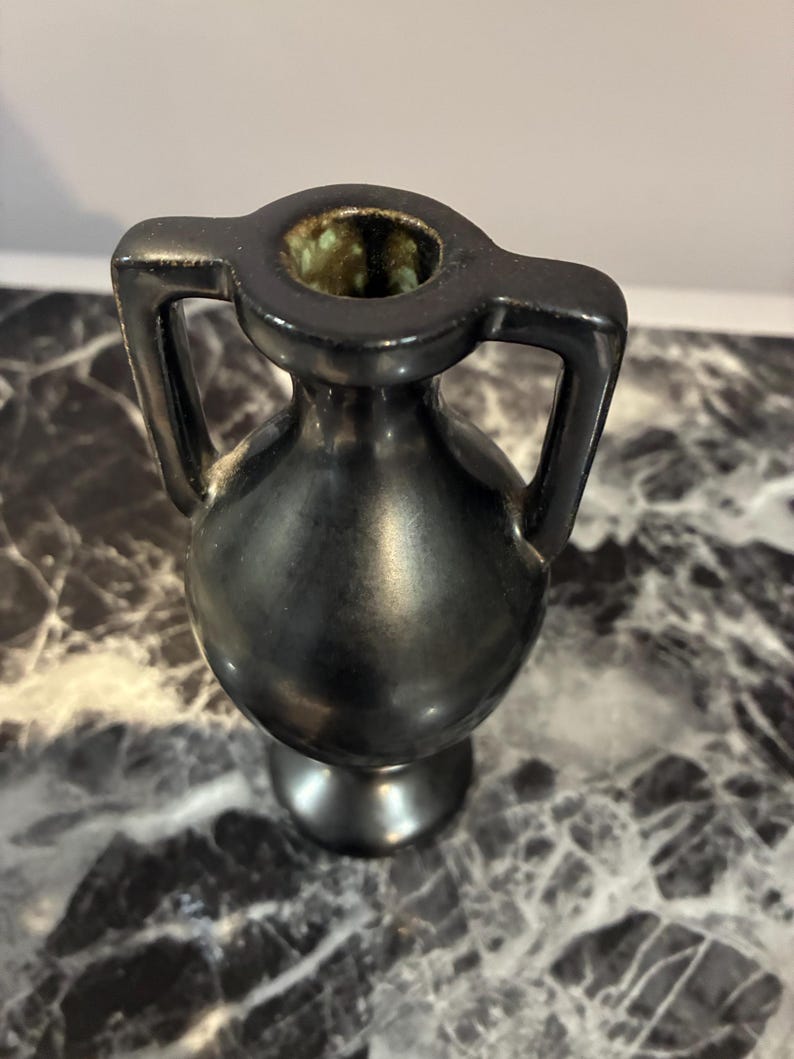 Pu&ograve; includere: Un piccolo vaso in ceramica nera con corpo arrotondato, collo stretto e due manici simmetrici che si estendono dal bordo alla spalla. L'interno dell'apertura presenta uno smalto maculato verde e marrone, in contrasto con l'esterno nero liscio.