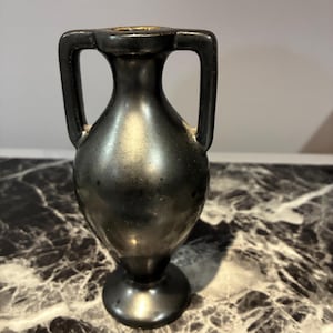 Pu&ograve; includere: Un piccolo vaso in stile anfora di colore nero metallizzato con collo stretto, corpo arrotondato e due manici verticali simmetrici che si estendono dal bordo alla spalla. Il vaso presenta una finitura liscia e riflettente e una base circolare.