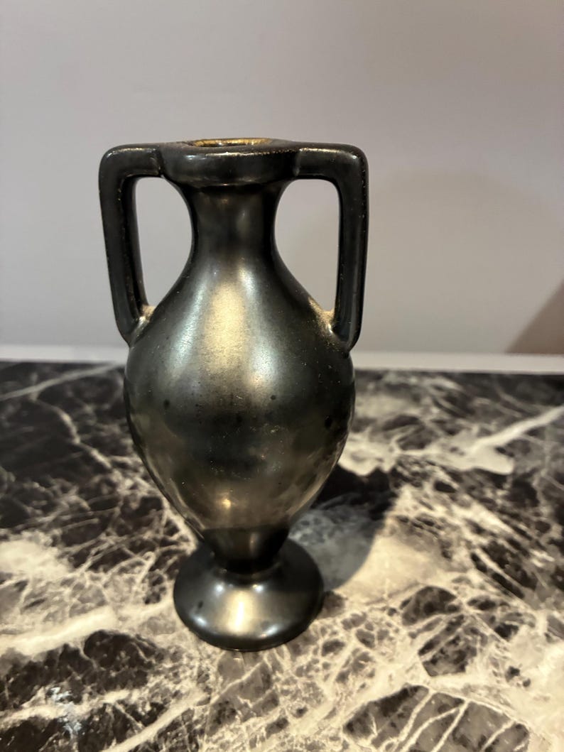 Pu&ograve; includere: Un piccolo vaso in stile anfora di colore nero metallizzato con collo stretto, corpo arrotondato e due manici verticali simmetrici che si estendono dal bordo alla spalla. Il vaso presenta una finitura liscia e riflettente e una base circolare.