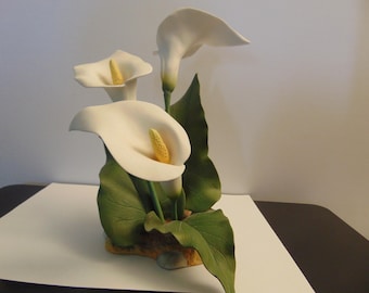 May include: A decorative sculpture featuring three white calla lily flowers with yellow spadices. The flowers are surrounded by large, textured green leaves that extend from a brown, rock-like base. The entire piece is crafted with smooth curves.