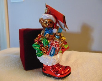 May include: A glass Christmas ornament shaped like a red boot filled with colorful wrapped gifts and a brown teddy bear wearing a Santa hat. The bear has a blue bow tie, and the boot features a white glittery cuff. A red tag is attached to the top.