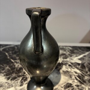 Pu&ograve; includere: Un piccolo vaso in metallo scuro con una finitura liscia e riflettente. Il recipiente presenta un collo stretto, un corpo arrotondato, un unico manico verticale che si estende dal bordo alla spalla e una base circolare.