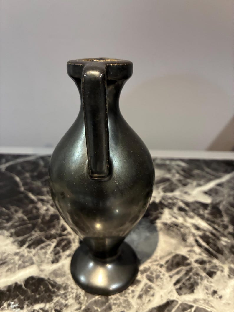 Pu&ograve; includere: Un piccolo vaso in metallo scuro con una finitura liscia e riflettente. Il recipiente presenta un collo stretto, un corpo arrotondato, un unico manico verticale che si estende dal bordo alla spalla e una base circolare.