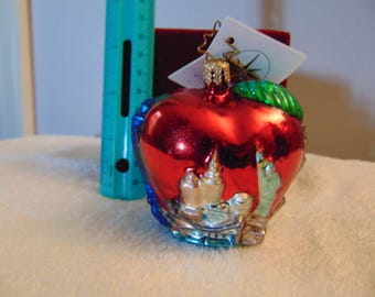 May include: A shiny red apple-shaped glass ornament featuring a metallic finish, a green leaf, and a gold-toned cap. A teal ruler is positioned next to the ornament, showing it stands approximately 3 inches tall. A white tag is attached.