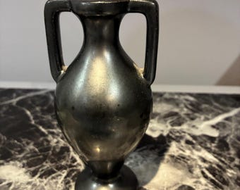 May include: A small, metallic black amphora-style vase featuring a narrow neck, a rounded body, and two symmetrical vertical handles extending from the neck to the shoulder, set upon a circular pedestal base with a smooth, reflective finish.