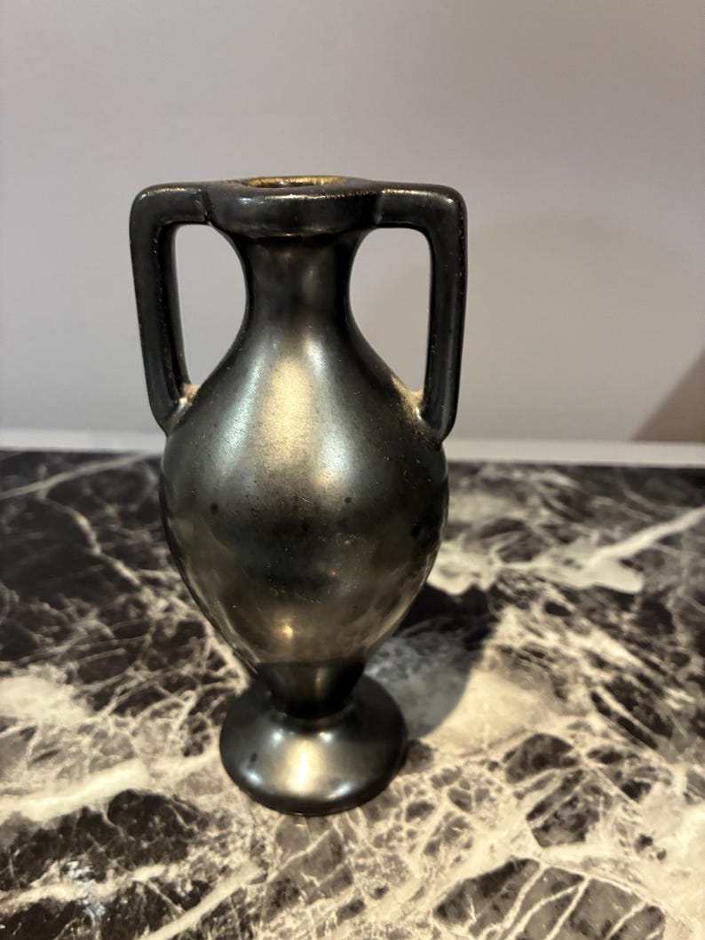 Pu&ograve; includere: Un piccolo vaso in stile anfora in metallo nero con collo stretto, corpo arrotondato e due manici verticali simmetrici che si estendono dal collo alla spalla, poggiante su una base circolare con una finitura liscia e riflettente.