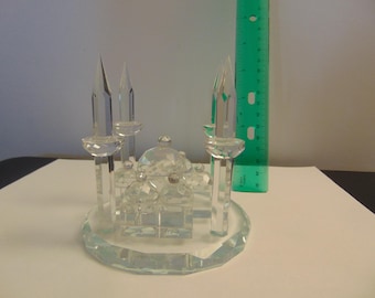May include: A clear glass figurine depicting a mosque with a central dome and four tall minarets on a circular base. A green plastic ruler is positioned vertically next to the object, showing the tallest minaret reaches approximately 3.5 inches.