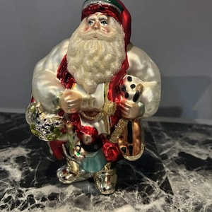 Könnte beinhalten: Ein Weihnachtsschmuck aus Glas in Form eines Weihnachtsmanns mit weißem, perlmuttfarbenem Mantel, roter Mütze mit grünem Rand und langem, weißem Glitzerbart. Er hält einen goldfarbenen Sack und ein kleines Musikinstrument, mit goldener Kappe.