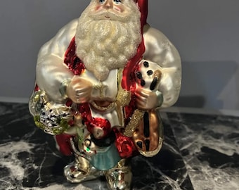 Può includere: Un addobbo natalizio in vetro a forma di Babbo Natale con cappotto bianco perlato, cappello rosso con bordo verde e una lunga barba bianca glitterata. Babbo Natale regge un sacco dorato e un piccolo strumento musicale, con tappo dorato.