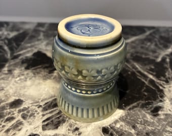 May include: A small, cylindrical ceramic vessel with a light blue and cream glaze. The upper section features a raised floral pattern, while the lower section displays a series of vertical stripes and a dotted band. The top rim is circular.