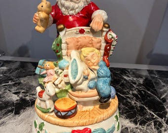 May include: A ceramic musical figurine featuring Santa Claus in a red suit holding a teddy bear, standing behind a sleeping child in a bed surrounded by toys including a doll, a rocking horse, a drum, and a blue stuffed animal on a base.