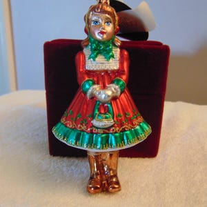 Könnte beinhalten: Ein Weihnachtsbaumschmuck aus Glas in Form einer Figur, die ein rotes Kleid mit grünen Verzierungen, einen grünen Schal und braune Stiefel trägt. Die Figur hält eine kleine silberne Handtasche. Der Saum ist mit goldenem Glitzer verziert.